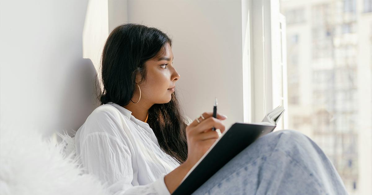 Woman sitting by a window writing in a notebook while thinking about how to support a friend experiencing an unexpected pregnancy.